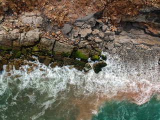 Aerial top view of sea waves hitting rocks on the beach with turquoise sea water. Amazing rock cliff seascape in the Portuguese coastline.