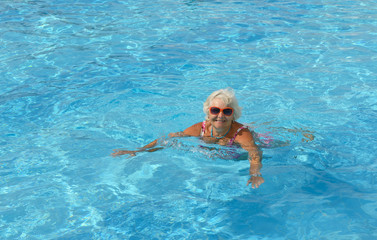 Aged woman is swimming in bright blue water of pool.