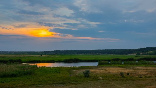 Timelapse sunset over river with reeds and forest under blue clouds