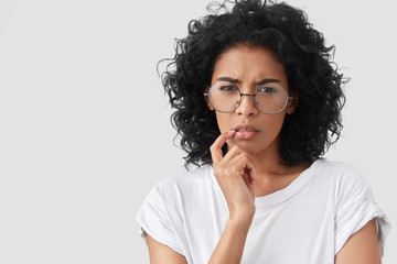 Fototapeta na wymiar Close up portrait of pretty serious displeased African American female frowns face, tries to concentrate and remeber something, wears round glasses and white t shirt, isolated on studio background