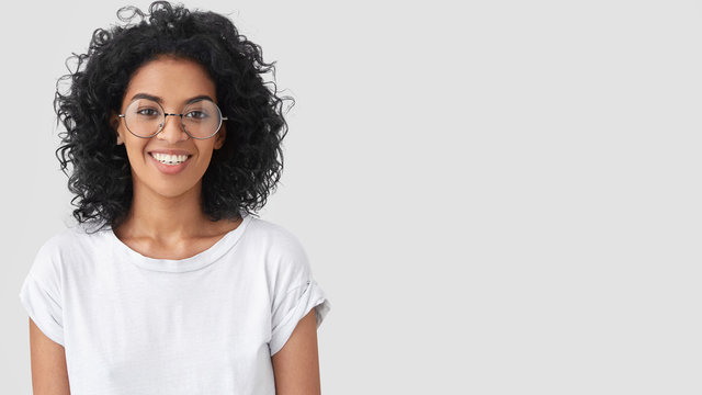 Positive African American female journalist has broad smile, curly bushy hair and dark healthy skin, satisfied after successful interview with celebrity, dressed in casual white t shirt, copy space