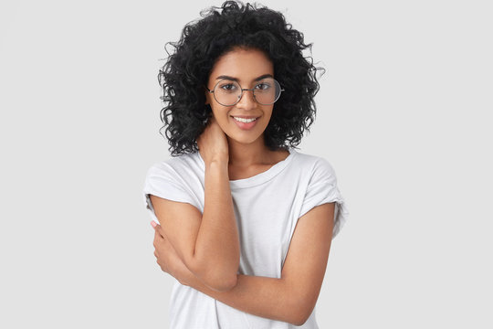 Horizontal shot of shy young mixed race woman with dark skin, curly hair, looks positively, keeps hand on neck and half crossed, happy to recieve help from handsome man, isolated over white background