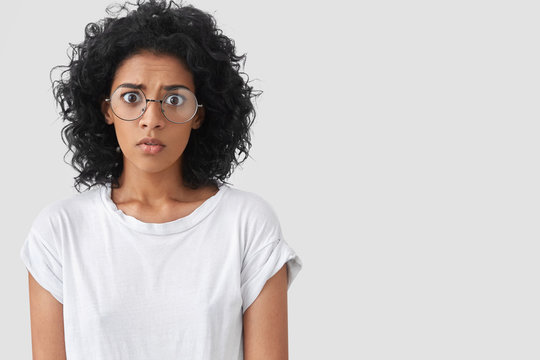 Frightened stupefied frustrated African American female looks in bewilderment with wide opened eyes, afraids of something, wears big spectacles, isolated over white background with copy space.