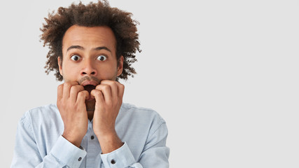 Studio shot of embarrassed mixed race male with nervous expression, bites finger nails, worries before interview, has scared expression, Afro hairstyle, poses against white blank wall with free space