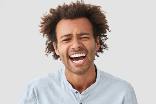 Optimistic glad African American male has broad smile, laughs at funny story, expresses positive emotions, being in good mood, isolated on white concrete studio wall. Happy mixed race young man