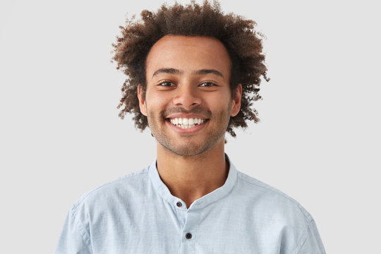 Headshot of handsome mixed race young man with shining smile, looks positively at camera, has white perfect teeth, stands against white background. People, lifestyle, emotions and fun concept