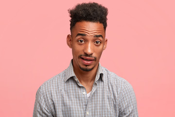 Confused attractive dark skinned male raises eyebrows in bewilderment, expresses surprisement, has beard and moustache, dressed in checkered shirt, isolated over pink background. Facial expressions