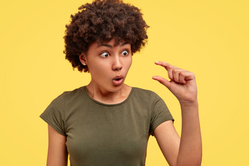 Surprised beautiful African American female stares with bugged eyes and makes tiny gesture, shows how much attention she recieves from boyfriend, poses against yellow background. It`s too small - obrazy, fototapety, plakaty