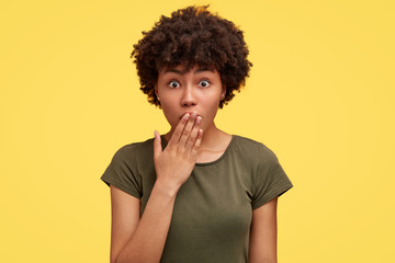 Studio shot of stupefied atrractive female closes mouth with palm, has bugged eyes and Afro hairdo, wonders latest news in world, isolated over yellow background. People and reaction concept