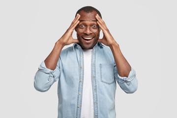 Portrait of cheerful young African American male giggles positively, rejoices good news, keeps hand on head, dressed in denim fashionable jacket, glad to be promoted at work. Positive emotions