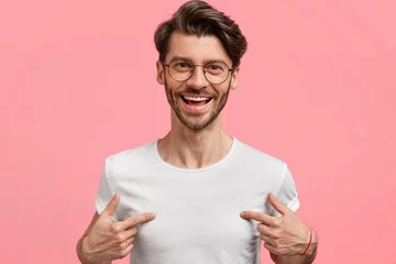 Fotobehang Horizontal shot of handsome cheerful hipster male indicates at white t shirt, shows blank space for your logo, being in high spirit, isolated over pink blank wall. Positive emotions and advertisement © Wayhome Studio