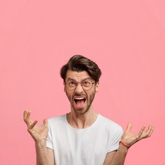 Aggressive attractive male with annoyed exppression, gestures angrily, yells, dressed in casual white t shirt, screams at friend, looks furious isolated on pink background. Negative emotions