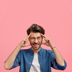 Vertical shot of stressed European male has headache after loud party, keeps hands on temples, dressed in denim shirt, has discontent expression, poses against pink background with blank space