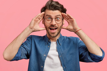 Overjoyed surprised young Caucasian male with positive surprised expression, keeps hands on head,...