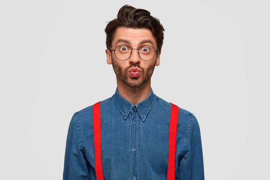 Handsome bearded male with trendy hairdo, keeps lips in kiss, makes grimace, dressed in fashionable outfit, isolated over white background. Attractive young hipster flirts with girlfriend indoor