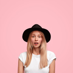 Vertical shot of attractive Caucasian young female with amazed worried expression, keeps mouth slightly opened, dressed in casual t shirt, poses against pink background with copy space upwards