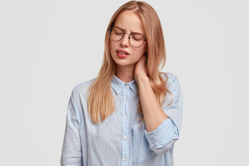 Tired overworked female has pain in neck, feels frustrated after long work at office, has fatigue expression, dressed in fashionable shirt, poses against white background. Tiredness concept.