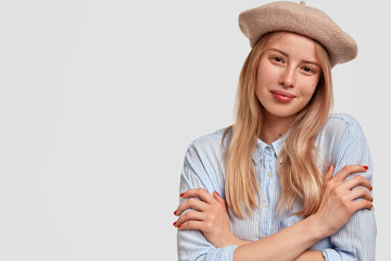 Lovely young female model with pleased expression, keeps arms folded, wears elegant beret, poses for fashion magazine, advertises fashionable clothes, isolated over white background with blank space