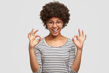 Studio shot of pleased young beautiful African American female shows ok sign, dressed in striped casual jacket, being in good mood, poses against white background. People and approval concept