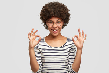Studio shot of pleased young beautiful African American female shows ok sign, dressed in striped...