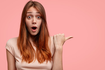 Horizontal shot of pretty young woman has amazed expression, freckles on face, being terrified with high prices in boutique, indicates at blank space with thumb, drops jaw and has bugged eyes