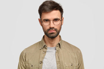 Headshot of handsome bearded serious young male freelancer with appealing appearance, looks directly at camera, dressed in fashionable shirt, works distantly, isolated over white background.