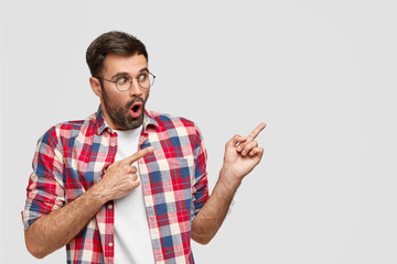 Horizontal shot of frightened shocked Caucasian male points with both fore fingers aside, has...