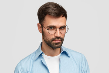Portrait of serious young male journalist wears round glasses and blue shirt, has confident look at camera, recieves important task from boss, listens attentively, isolated over white background.