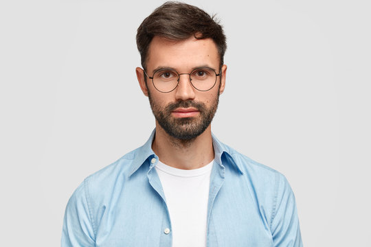 Headshot of attractive bearded young European male with serious confident expression, looks directly at camera, wears spectacles, isolated over white background, thinks about starting family life.