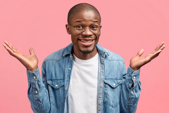 Positive dark skinned bald male raises palms in hesitation, dressed in fashionable denim jacket, can`t make decision where to travel, stands against pink background. Clueless African American man