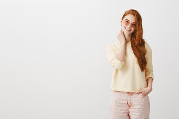 Obraz premium Shy girl talking with guy she likes. Portrait of attractive trendy woman with ginger hair touching neck and smiling broadly, being nervous and timid while standing against gray background