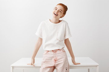 Woman waiting for friend dress up, leaning on table in room and talking casually, laughing and making jokes. Charming redhead girl with bun hairstyle, tilting head and smiling broadly at camera