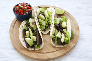 Corn tortillas with grilled beef, avocado, lime and salsa on wooden board, close-up. White wooden background. Side view.