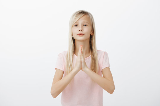 Portrait of charming adorable little kid with blond hair, holding hands in pray over chest and looking with angel expression at camera, asking parents favour or begging for candy, having big desire