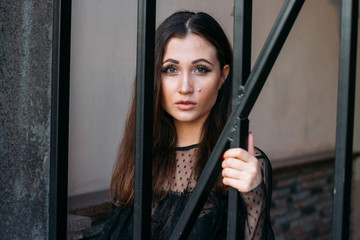 city portrait. sad girl. brunette in a black dress. expectation. dreams. portrait of a young, beautiful girl. The brunette in a black dress is standing near the cage with black rods. clings to them.