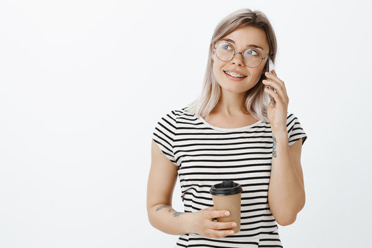 Come and pick me up in local cafe. Stylish relaxed good-looking woman with fair hair in glasses, holding cup of coffee, smiling and gazing aside while talking on smartphone, making plans for evening
