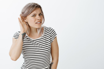 Repeat louder, cannot hear your whisper. Portrait of intense confused attractive female student in striped t-shirt, bending towards camera, holding hand near ear, gossiping or eavesdropping