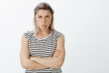 I do not want talk with you. Portrait of disobedient angry young woman with fair hair, sulking and frowning, sticking out tongue and holding hands crossed, being offended or insulted, arguing