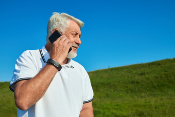 Sideview of sportsman talking on cellphone while sunny day. Wearing white polo shirt with dark blue stripes. Looking at side. Having sportwatch on left hand. Outdoors activities. Fresh air.