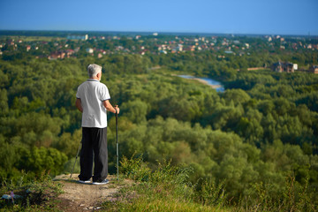 Old man watching beatiful city panorame keeping tracking sticks. Standing on green grassy hill. Wearing white polo shirt with dark blue stripes,black trousers, sneakers. Healthy lifestyle.