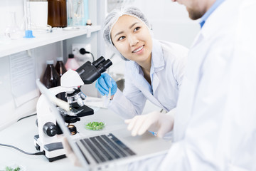 Crop view of smiling female microbiologist standing at eyepiece of microscope with samples of green...