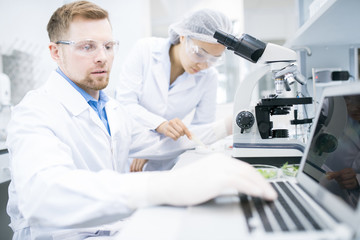 Side view of male microbiologist in white laboratory coat typing on laptop keyboard data received during testing of food nutrition properties under microscope with female assistant on background.