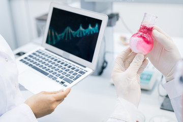 Crop hands of microbiologist holding flask with meat sample solution standing at colleague holding laptop with computer DNA model