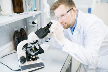 Crop view of man in glasses and laboratory coat standing at desk and looking at eyepiece of microscope with green vegetable sample lying on it