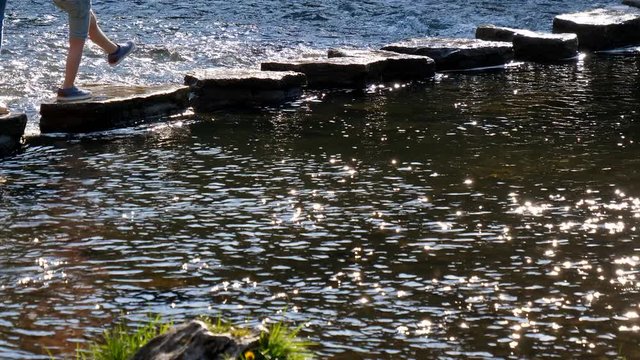 Legs of two mothers with children are crossing fast river walking stepping stones