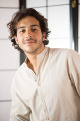 An handsome young man, 20Y, is posing in studio and smiling, with eyes half closed. White skirt. Retroilluminated booths in the background.