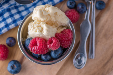 A scoop of vanilla ice cream with raspberries and blueberries on a wooden board and blue and white dish towel.
