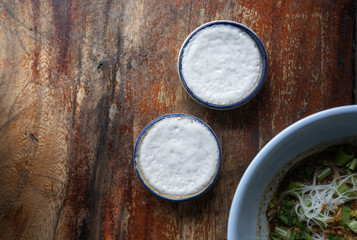 top view, khanom thuay , Thai dessert coconut milk custard in small porcelain cup ,with Thai Boat noodles on wooden background