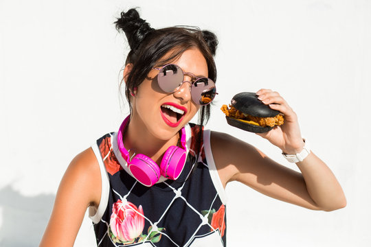 stylish sporty funny girl hipster with headphones around her neck, eat fast food black burger and french fries, white background, trend of the summer season