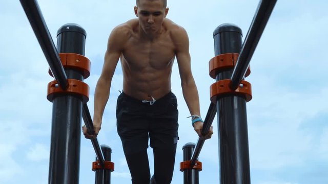 A lean and muscular guy trains on workout bars. In the background a cloudy sky.